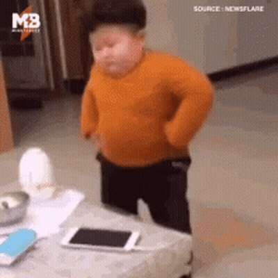 A Little Boy In An Orange Sweater Is Standing Next To A Table . GIF