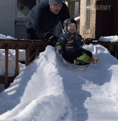 A Little Boy Is Sliding Down A Snowy Hill While A Man Stands Behind Him GIF