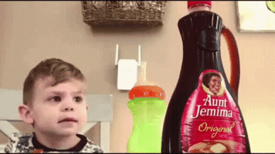 A Little Boy Is Standing In Front Of A Bottle Of Aunt Jemima Original Syrup GIF