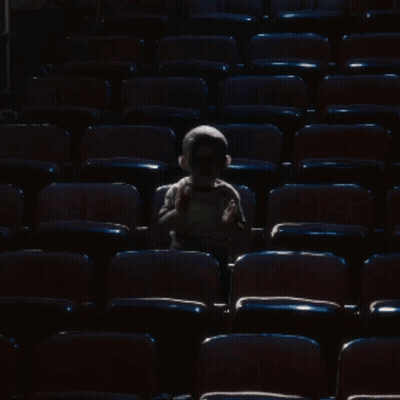 A Little Boy Sits In A Dark Auditorium With A Few Empty Seats GIF