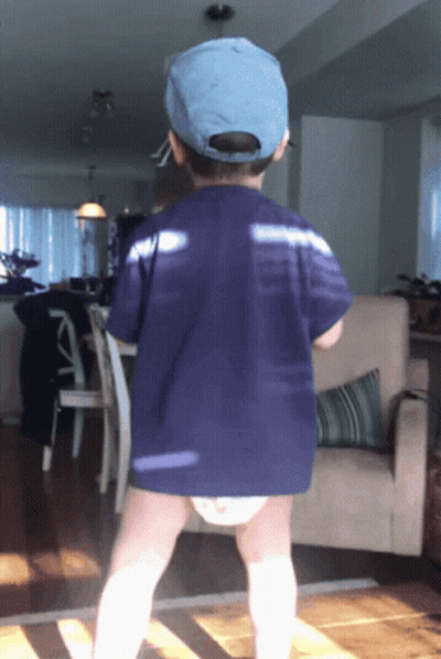 A Little Boy Wearing A Blue Hat And A Diaper Stands In A Living Room GIF