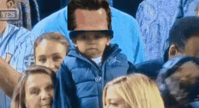 A Little Boy Wearing A Hat Is Sitting In A Crowd Of People GIF