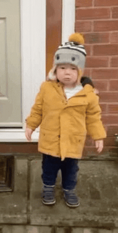 A Little Boy Wearing A Yellow Coat And A Hat Is Standing In Front Of A Brick Wall . GIF