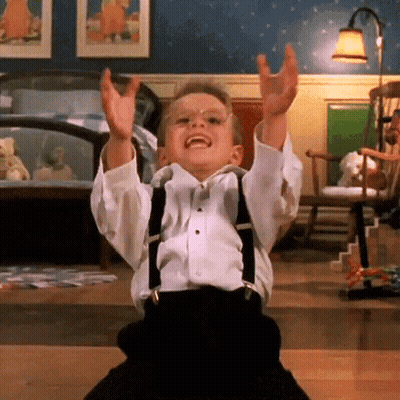 A Little Boy Wearing Suspenders And A Bow Tie Is Sitting On The Floor With His Arms In The Air GIF