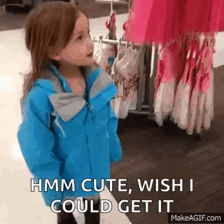 A Little Girl In A Blue Jacket Is Standing In Front Of A Rack Of Clothes And Talking To Someone . GIF