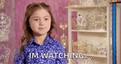 A Little Girl In A Blue Shirt Is Sitting In Front Of A Shelf With Trophies And Says `` Im Watching ... '' . GIF