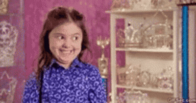A Little Girl In A Blue Shirt Is Smiling In Front Of A Shelf Full Of Trophies . GIF