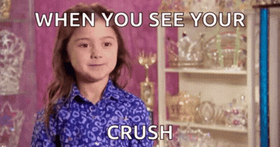 A Little Girl In A Blue Shirt Is Smiling While Standing In Front Of A Shelf With Trophies On It . GIF