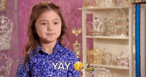 A Little Girl In A Blue Shirt Is Standing In Front Of A Shelf And Making A Funny Face . GIF