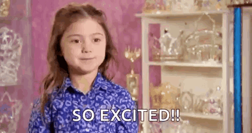 A Little Girl In A Blue Shirt Is Standing In Front Of A Shelf And Says `` So Excited ! '' GIF