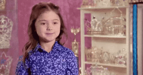 A Little Girl In A Blue Shirt Is Standing In Front Of A Shelf Filled With Tiaras . GIF