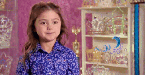 A Little Girl In A Blue Shirt Is Standing In Front Of A Shelf Filled With Tiaras And Trophies . GIF