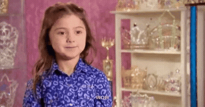 A Little Girl In A Blue Shirt Is Standing In Front Of A Shelf Full Of Trophies . GIF