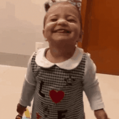 A Little Girl In A Dress With A Heart On It Is Smiling And Laughing . GIF