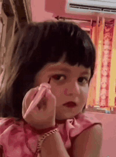 A Little Girl In A Pink Dress Is Applying Makeup To Her Face GIF
