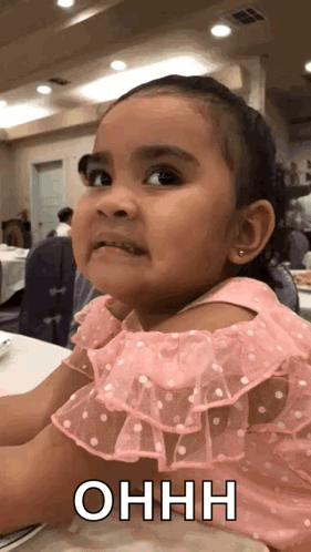 A Little Girl In A Pink Dress Is Sitting At A Table Making A Face . GIF