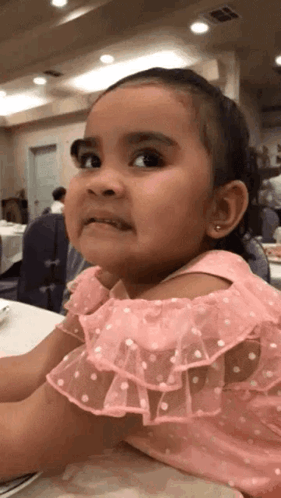 A Little Girl In A Pink Dress Is Sitting At A Table Making A Funny Face GIF