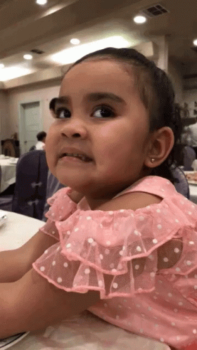 A Little Girl In A Pink Dress With White Polka Dots Is Sitting At A Table GIF