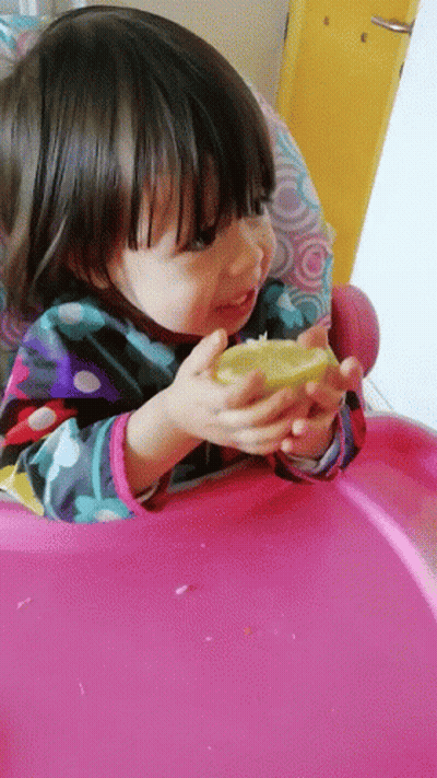 A Little Girl In A Pink High Chair Is Eating A Lemon GIF