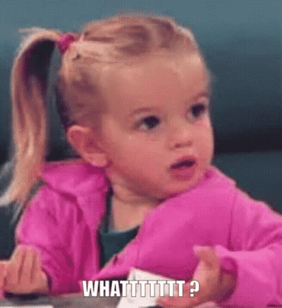 A Little Girl In A Pink Jacket Is Sitting At A Table Making A Funny Face And Saying What ? GIF