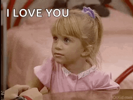 A Little Girl In A Pink Shirt Is Sitting At A Table With A Person And Says `` I Love You '' . GIF
