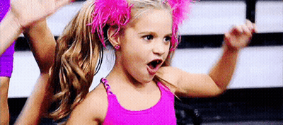 A Little Girl In A Pink Tank Top And Pink Feathers In Her Hair Is Making A Funny Face . GIF