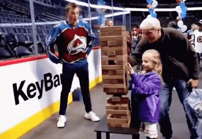 A Little Girl In A Purple Jacket Is Playing Jenga With A Man In A Hockey Jersey Behind Her GIF