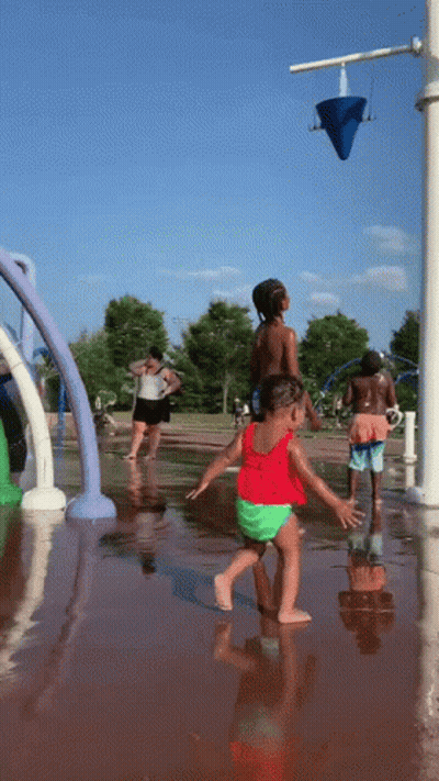 A Little Girl In A Red Top And Green Shorts Is Playing In A Water Fountain GIF