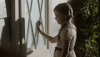 A Little Girl In A White Dress Is Looking At A Window GIF
