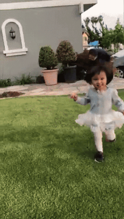 A Little Girl In A White Dress Is Running On The Grass In Front Of A House GIF