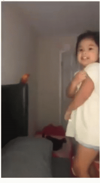 A Little Girl In A White Shirt Is Standing In A Room And Smiling GIF