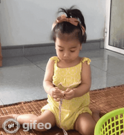 A Little Girl In A Yellow Dress Is Sitting On The Floor Playing With A Ribbon And A Basket With Gif Written On It GIF