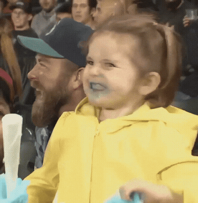 A Little Girl In A Yellow Jacket Is Making A Funny Face In A Crowd GIF