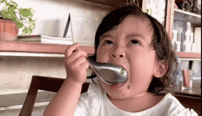 A Little Girl Is Eating Something With A Spoon In Her Mouth GIF