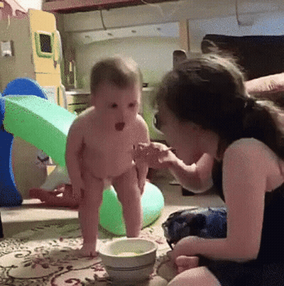 A Little Girl Is Feeding A Baby From A Bowl On The Floor GIF