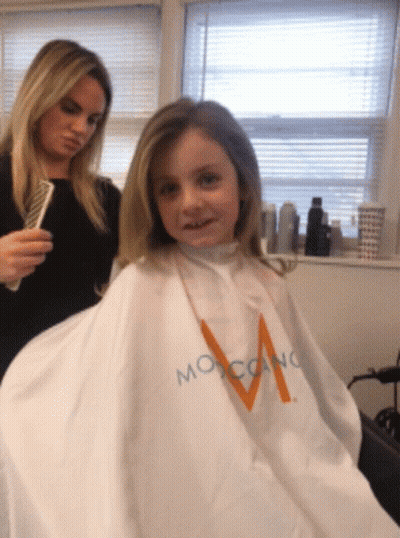 A Little Girl Is Getting Her Hair Cut By A Woman Wearing A White Cape That Says Moroccano GIF