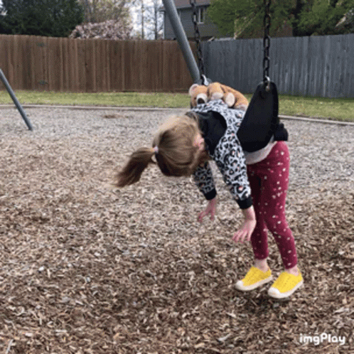 A Little Girl Is Hanging Upside Down On A Swing In A Park GIF