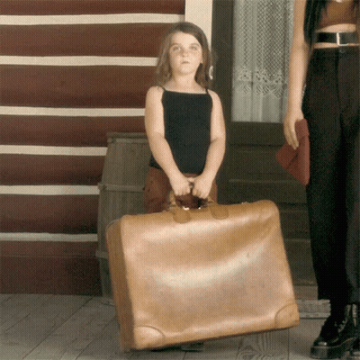 A Little Girl Is Holding A Large Brown Suitcase In Front Of A Log Cabin GIF