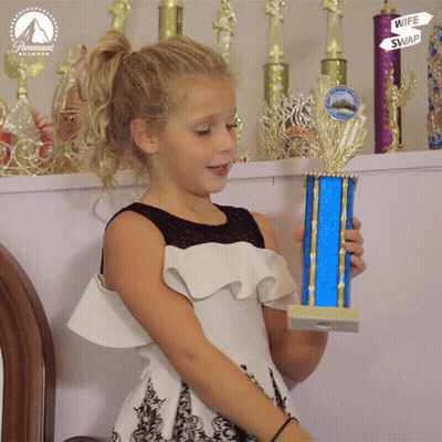 A Little Girl Is Holding A Trophy In Front Of Trophies That Say Wife Swap GIF