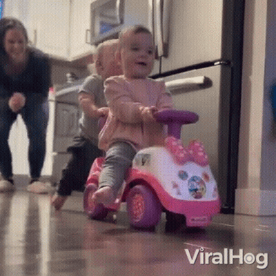 A Little Girl Is Riding A Pink And White Toy Car While A Little Boy Watches . GIF