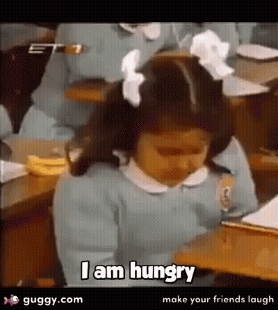 A Little Girl Is Sitting At A Desk Saying I Am Hungry . GIF