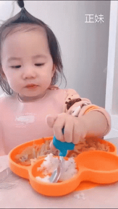 A Little Girl Is Sitting At A Table With A Plate Of Food And A Spoon In Her Hand . GIF