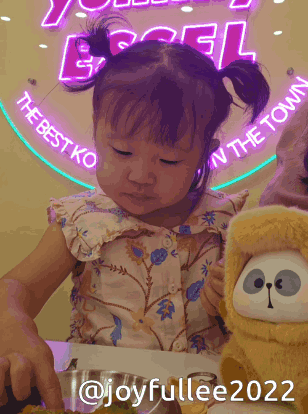 A Little Girl Is Sitting At A Table With A Stuffed Animal In Front Of A Sign That Says The Best Ko In The Town GIF