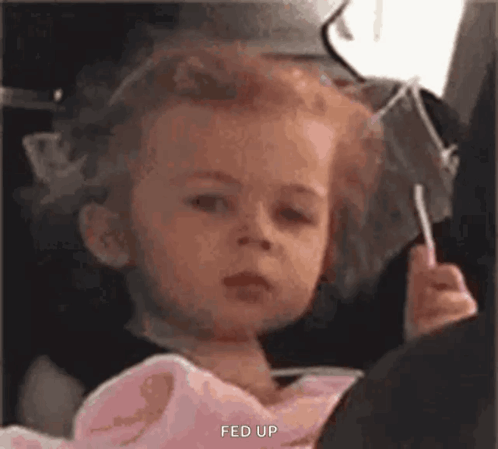 A Little Girl Is Sitting In A Car Seat Holding A Lollipop And Making A Funny Face . GIF