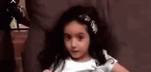 A Little Girl Is Sitting In A Chair With A Flower In Her Hair And Making A Funny Face . GIF