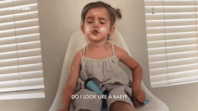 A Little Girl Is Sitting In A Chair With Her Eyes Closed And The Words Do I Look Like A Baby Above Her GIF