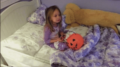 A Little Girl Is Sitting On A Bed Holding A Pumpkin Bucket GIF