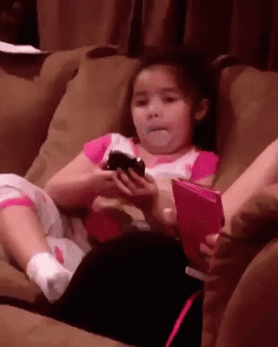 A Little Girl Is Sitting On A Couch Holding A Cell Phone And A Book . GIF