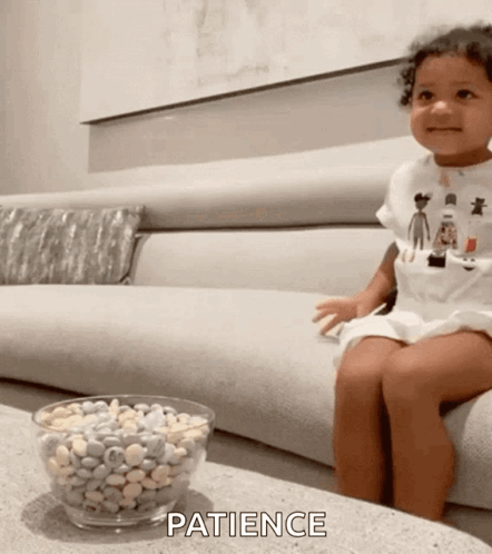 A Little Girl Is Sitting On A Couch Next To A Bowl Of Candy And The Word Patience Is On The Table . GIF