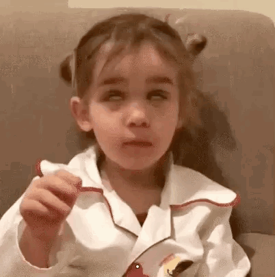 A Little Girl Is Sitting On A Couch With Her Eyes Closed And Eating A Cookie . GIF
